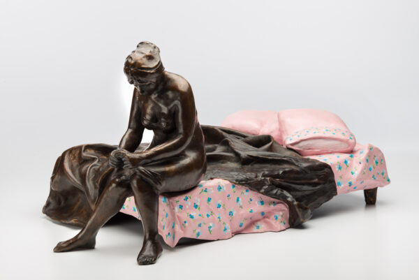 A woman sits at the end of an unmade bed.