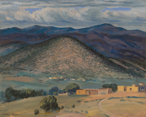 Adobe buildings are at the base of a hill with mountains in the background.