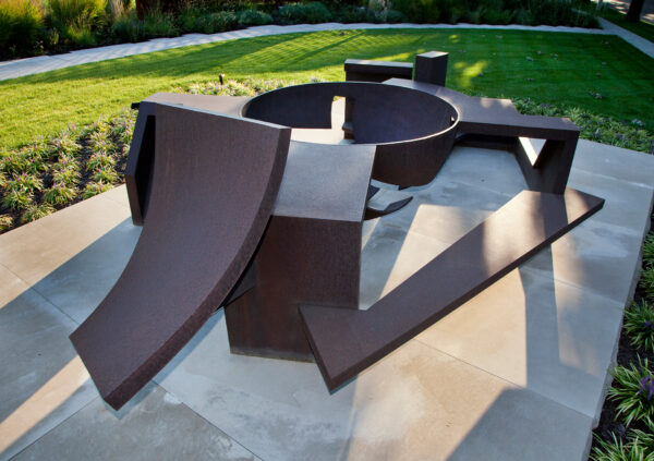 A sculpture of abstracted shapes. There are six pieces bolted together. At the center is a large bowl shape. On one end is a curved shape and the rest of the sculpture is flat step shapes.