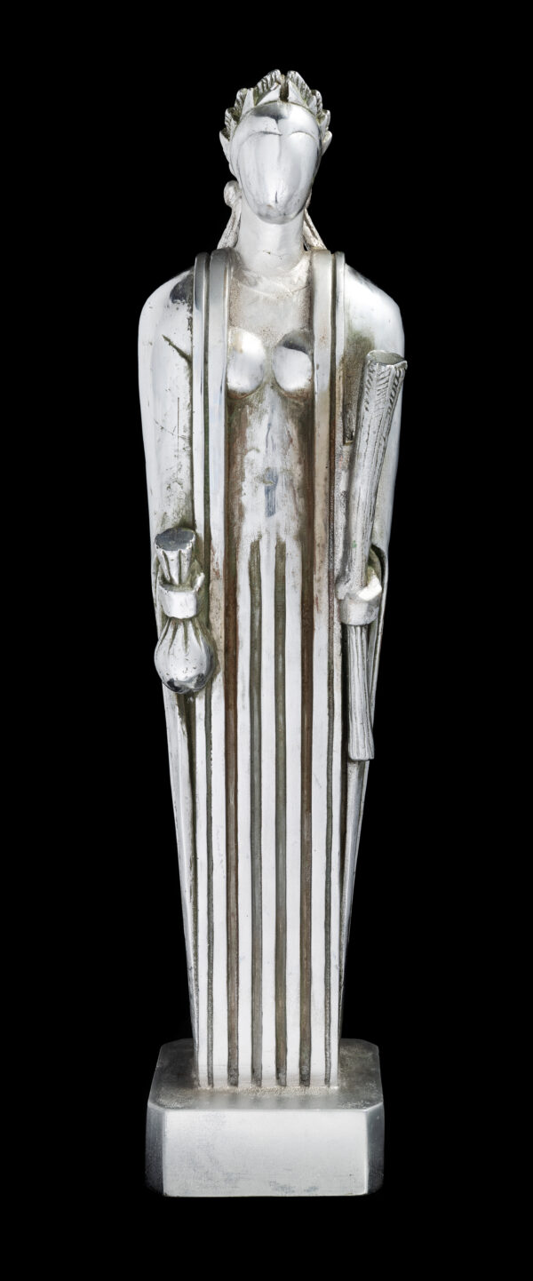 A maquette of silver-looking abstract sculpture of a woman with no face, wearing a crown and long robes with folds of fabric. She holds a bag in her right hand and a swath of wheat in her left hand