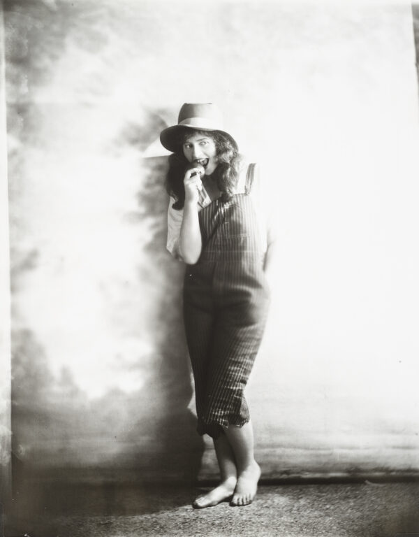 A woman in overalls and straw hat stands in front of a blank wall or canvas.