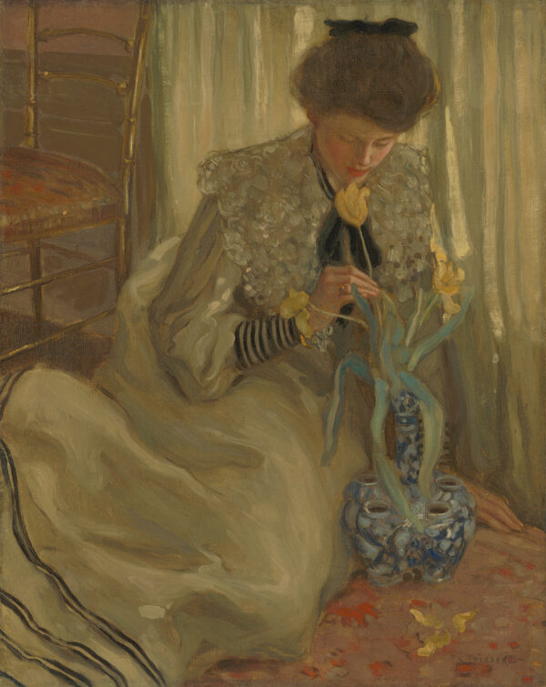 A young woman is seated on the floor leaning forward to admire one of three yellow tulips that extend from a blue and white Delftware bulb vase.