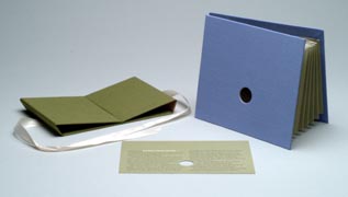 Accordion-style folded paper between two cardboard and fabric end pieces (a) expands to reveal a three-dimensional photographic image of a figure falling into a well (design based on Dante's Inferno) when viewed through an opening (peephole) in either end; piece has a green cardboard and fabric folding cover (b) that ties with ribbon and includes an information card (c).