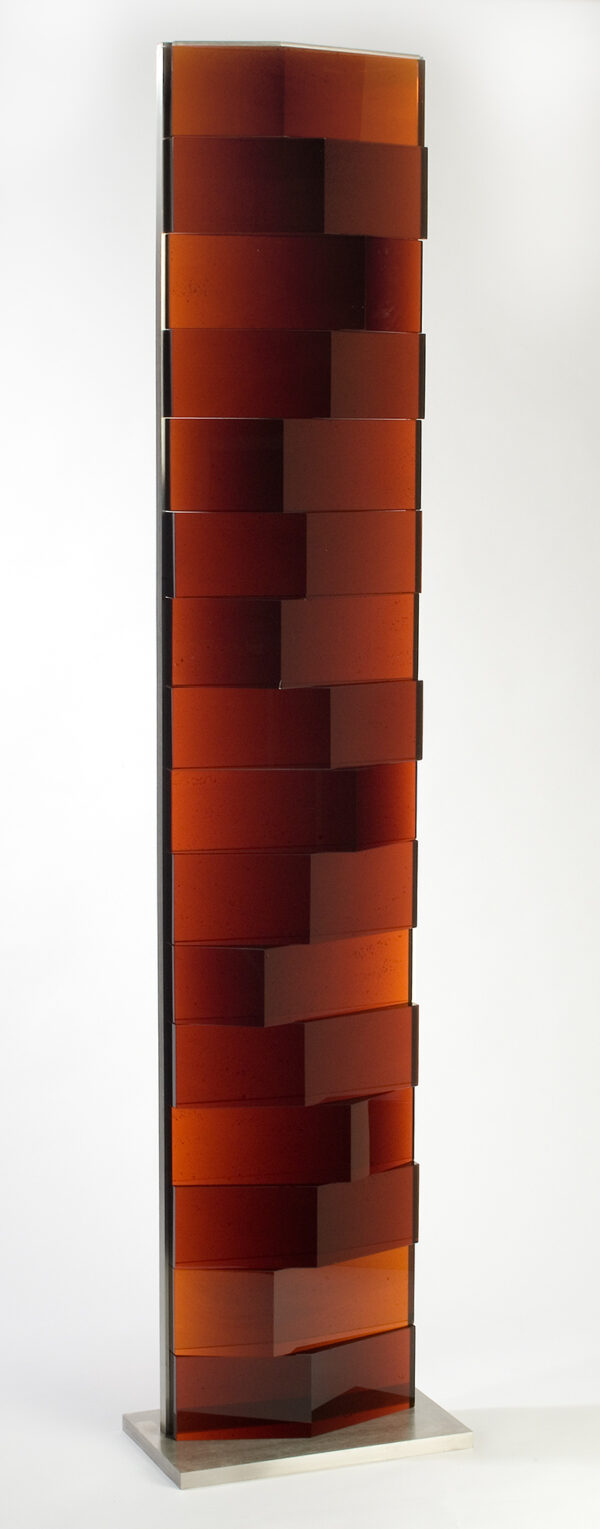 The sculpture has a stainless steel rectangular base from which vertical supports hold blocks of dark red glass stacked one on top of another. The blocks of glass are rectangular with a triangular projection that varies in size at the front of each block.
