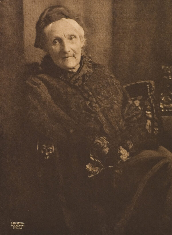 A portrait of a seated woman. She wears a dark wig over her white hair. Her hands are crossed on her lap but can’t be seen.