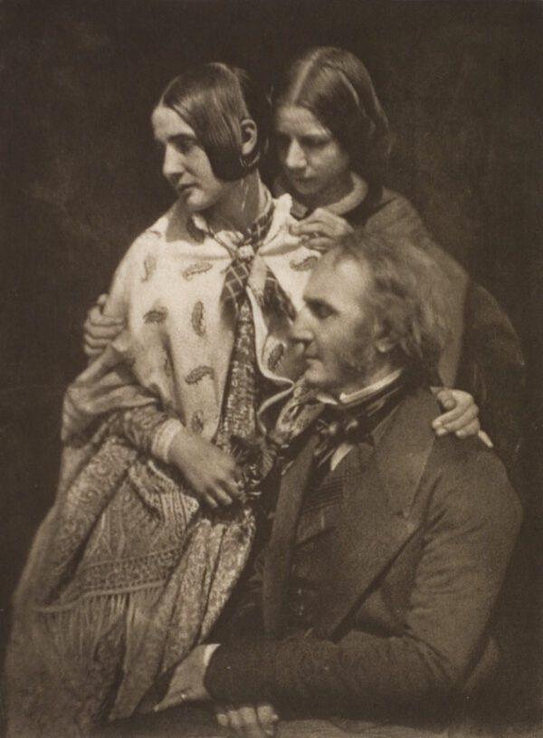 A group portrait of two women standing beside a man sitting. The man’s hands are folded on his lap. The woman in front has her left arm around the man and the woman in back has both her hands around the woman in front.