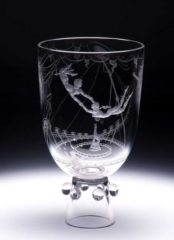 Shape # X2623. A vase standing on a narrow foot that includes round balls circling the foot. The engraved scene fully circles the vase with a circus that includes three “aerialists”, the big top tent, a juggling clown and seal with ball.