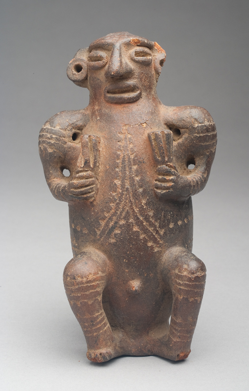 The figure has a red body clay with dark brown surface. He is holding something in his hands. Mallets? Fans? His has incised decoration throughout. He has a rattle inside.