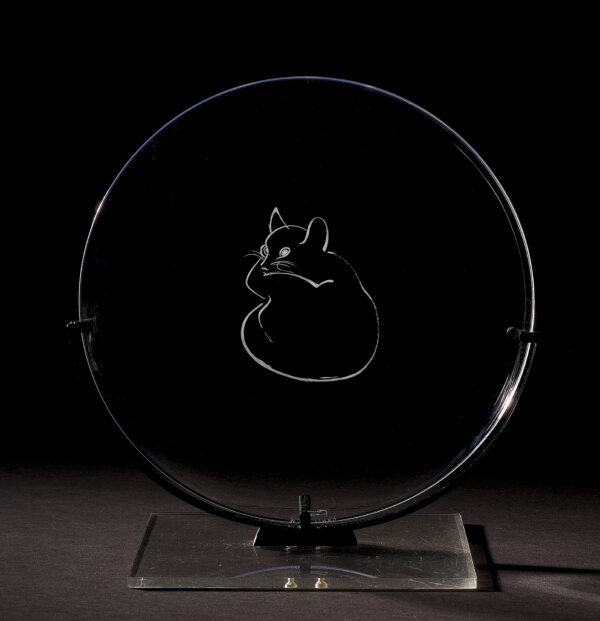 A clear glass plate with the image of a curled-up cat engraved on the center. The plate includes a holder of a plexi and wood base with metal arms painted black.