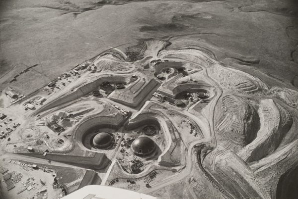 Round missile silos surrounded by banked earth and rolling empty hills in the background. The tip of the plane wing is visible at lower edge.