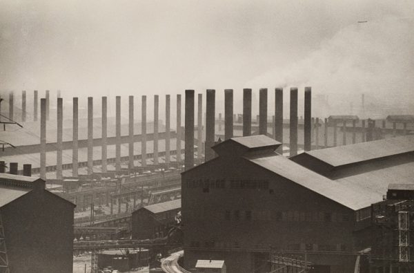 Scene of a steel mill with buildings and smoke stacks.