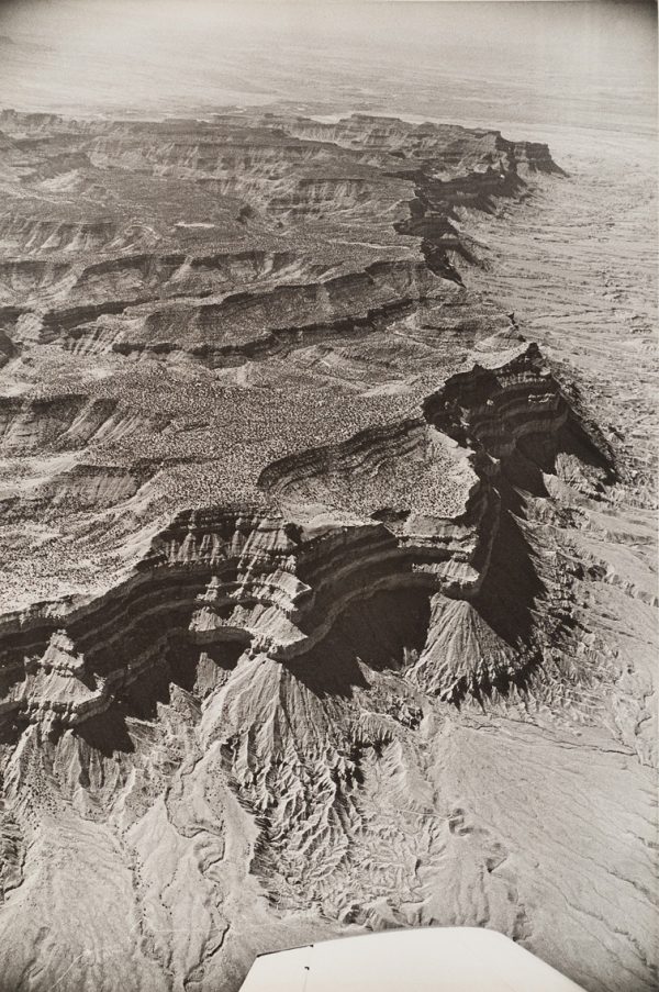 A view of cliffs in an arid landscape with plains in the distance. The plane’s wing is visible at bottom center.