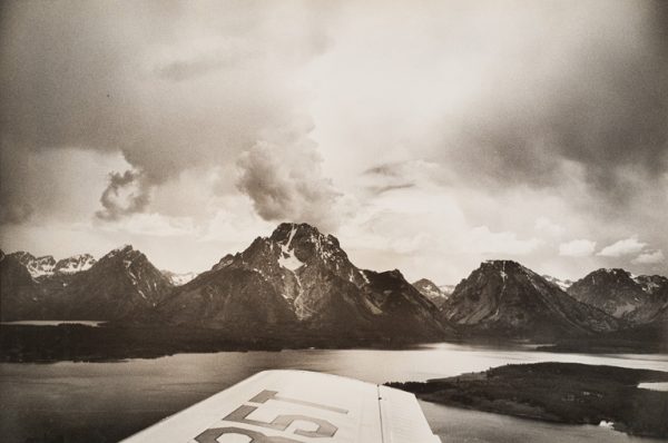 Lake, mountains with snow and clouds. Plane’s wing is visible at lower center.