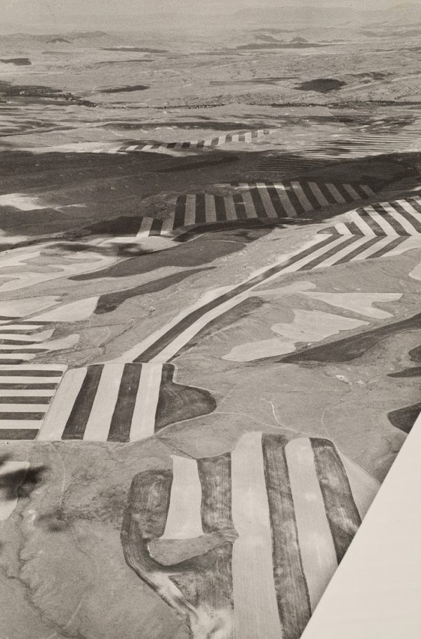 Patch work of fields with strong dark/light stripes. Wing of plane can be seen in lower right corner. Clouds are casting a shadow near the top.