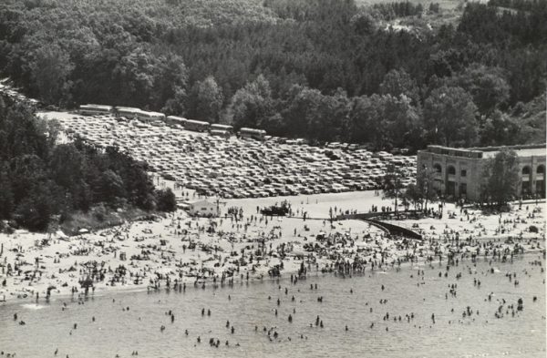 Water and a beach in the foreground that is full of people. A building is on the right and a full parking lot in the top half of the photo.