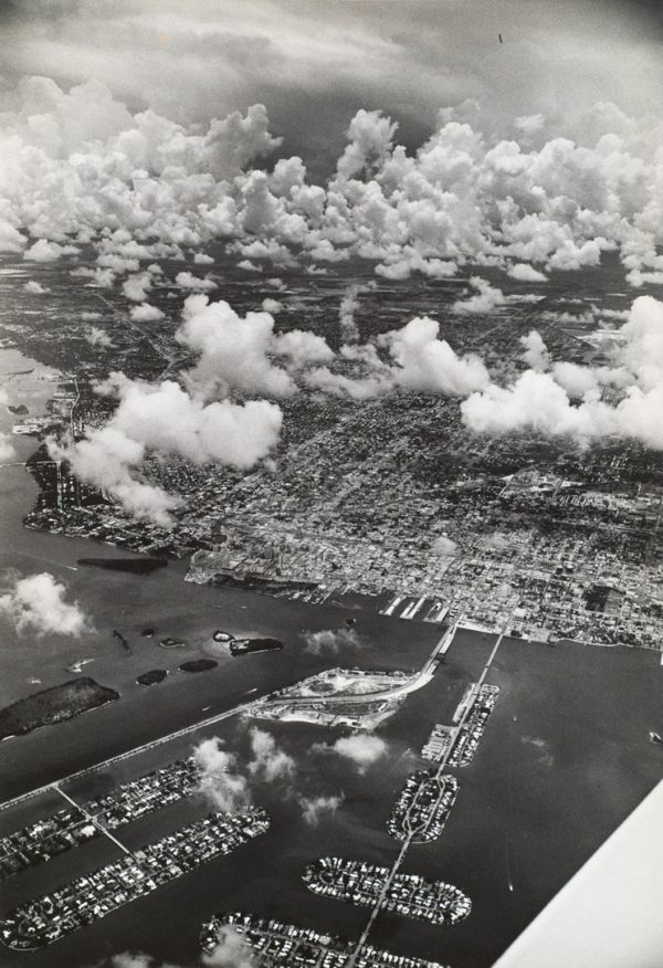 A view of Miami with water and piers in foreground and the city half obscured by fluffy clouds in the background.
