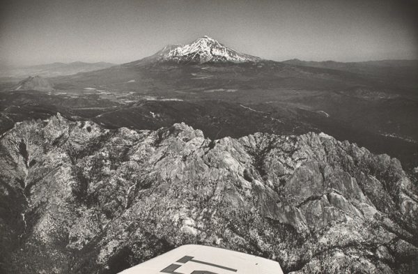 Mt. Shasta is seen in the distance with rough hills in front and the plane’s wing visible at bottom center.
