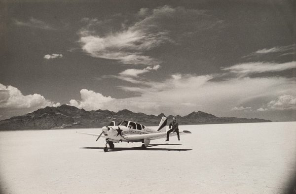 A small plane with a man sitting on one wing is in a desert with mountains in the background.