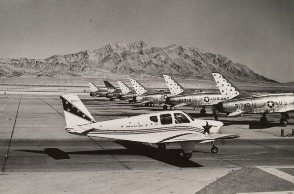 View of airfield with 7 airplanes on the tarmac and mountains beyond.