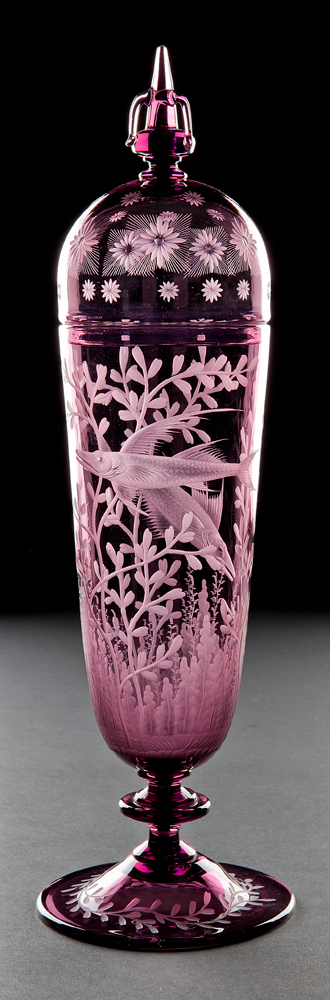Steuben # 6847 a form derived from 16th century Venetian glass. Tall vase shape with round cover terminating with a conical shape and three pulled arches. The engraving is a marine environment of two fish swimming in a variety of sea grasses. The foot has a wreath of stylized vines and leaves and the cover has a variety of star burst designs.