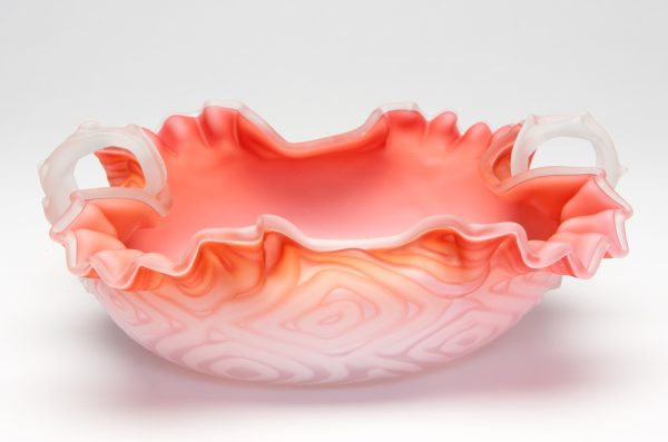 A shallow bowl with coral color on the inside gradient to white on the bottom. It is cased in white and clear glass outside which has air trapped in a geometric pattern. There are two small handles in a leaf shape.