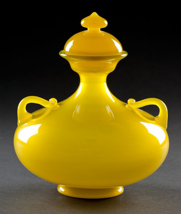 A yellow-green slightly opaque blown glass vase. There are two small handles on a rounded base that narrows to a small mouth. With domed, pointed cover.