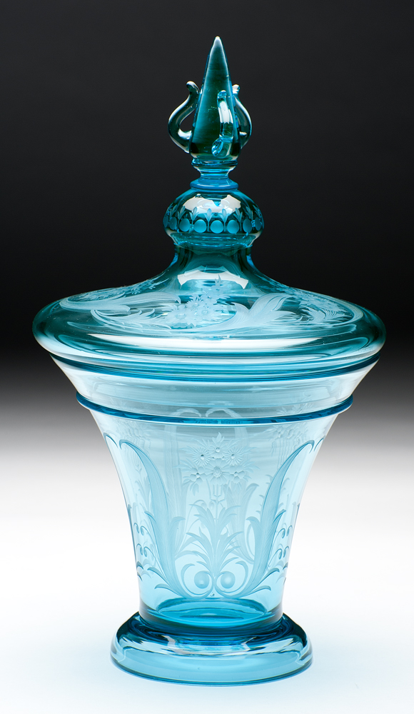 A covered vase in “Marina blue” colored glass. The sides and top of the cover are etched with floral designs. The cover has a pointed finial.