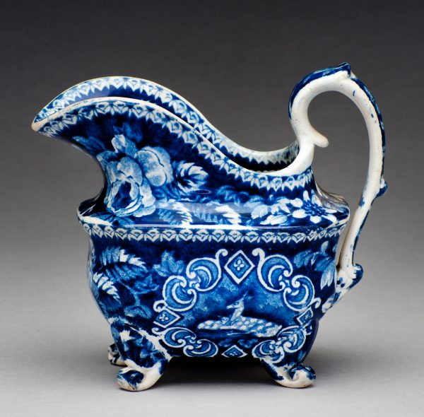 A blue transfer of a deer and doe in a frame of geometric designs and flowers. The creamer is on four scroll feet.