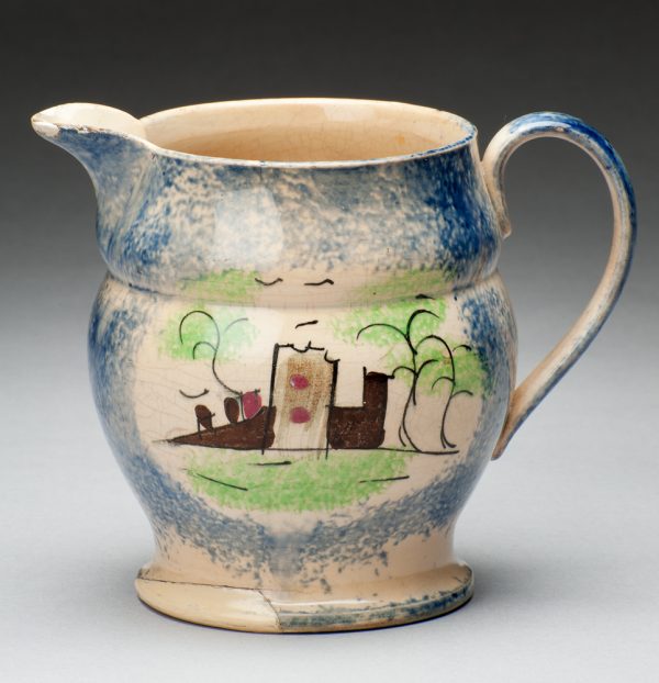 Spatterware creamer in the Fort pattern, blue with scene of buildings. The opposite side of the creamer has a scene of grass, tree and fence.