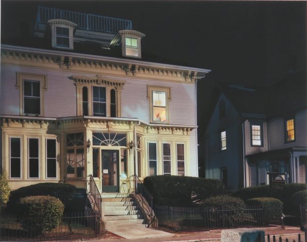 A white house is framed by the black night. A street light strikes the lower half of the house. There are interior lights in the top right attic window and the second floor right window. A woman can be seen in that window.