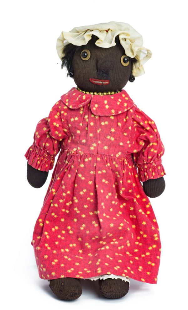 A female doll in a red dress printed with yellow flowers. She has a white bonnet and a gold necklace. Her mouth is embroidered, with solid button eyes and ears. Her hair is faux fur.
