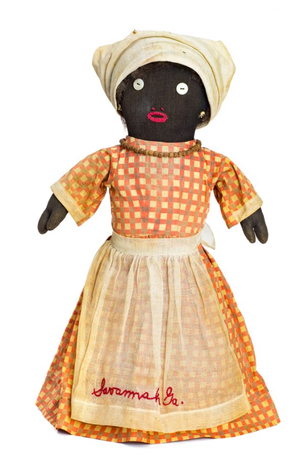 A female doll, wears a red plaid dress, white scarf and apron, and bead necklace. On the apron is embroidered 