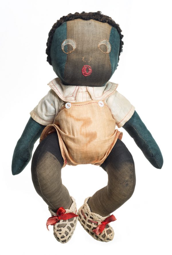 The baby doll wears a tan jumper, white shirt and crocheted booties with red bows. The baby’s features are embroidered.