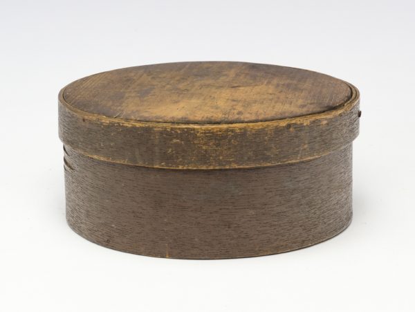 A small round lidded box made from bent wood. The exterior of the box and lid were painted brown.