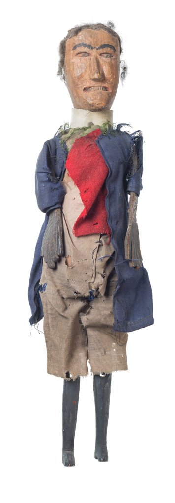 A male doll made of carved and painted wood dressed in cotton and wool clothing. The man is wearing high waisted khaki pants with a blue overcoat. A red wool cloth and green fabric cover his chest. An off-white plastic collar runs around his neck and is secured with a nail: this possibly indicates that he is a clergy man.