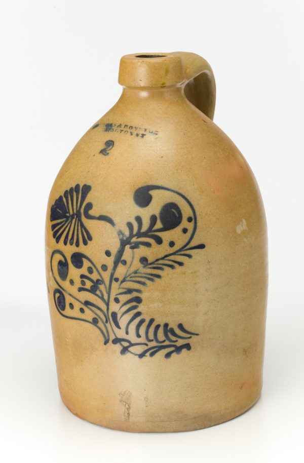 A two-gallon tan jug with fancy cobalt leaf pattern on side. The jug has a lug handle.
