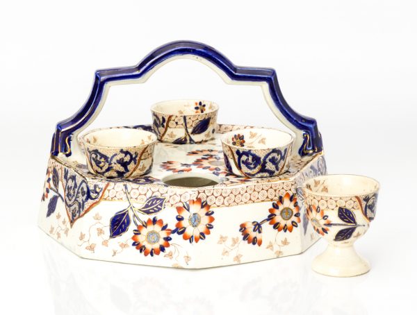 Mold-made ceramic caddy and egg cups with a white clay body and several layers of decorative surface: underglaze painting in blue, printed decoration in orange, clear glaze, and over-glaze painting in orange and gold.