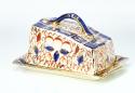 A butter dish with a lid that is on a slant of blue, red and gold decorations over cream base