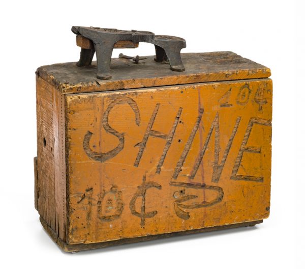 An orange box with a cast metal footrest, a place to put your shoe to be shinned. The box has a door to the inside where supplies could be kept.