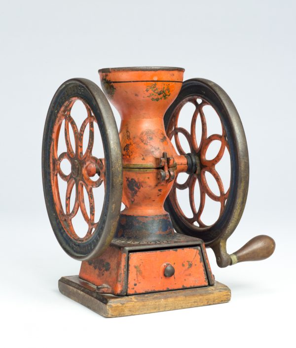 A coffee grinder composed of an hour glass shaped body with two ornate wheels. The grinder was made from assembled cast iron and steel elements, with the exterior painted red with applied decal designs of foliate in dark red, green, and black. The grinder has a wood handle and is mounted to a wood base.