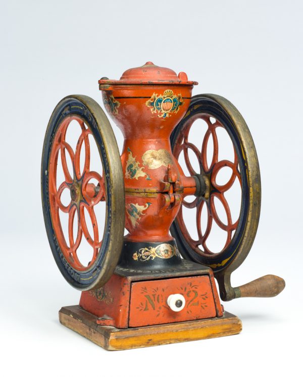 A coffee grinder composed of an hour glass shaped body with two ornate wheels. The grinder was made from assembled cast iron and steel elements, with the exterior painted red with applied decal design. The grinder has a wood handle, a metal drawer to collect the grounds, and is mounted to a wood base. A ceramic knob acts as the drawer pull.