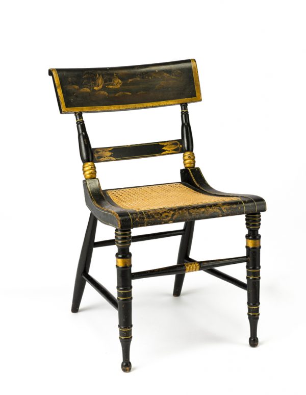 Chairs are ebonized with gold stencils and cane seats. The back has a scene of two ships in a cove, with trees to left, rocks around the shore in the foreground, and a house with lights in the windows at the right. The lower back rail has stencils of stylized leaves. The front of the seat also has stenciled pattern.