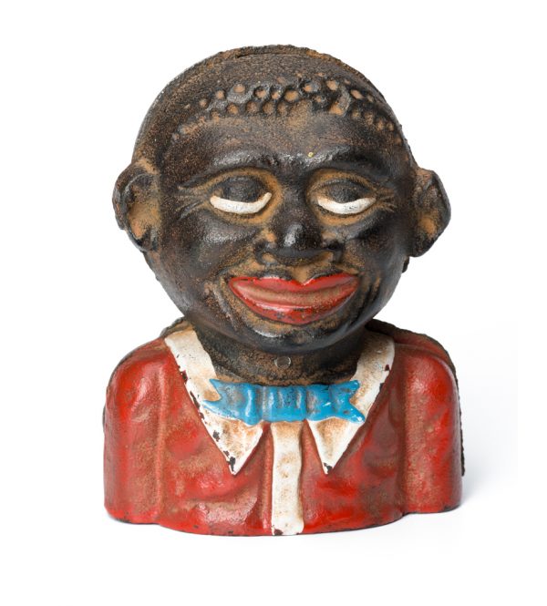 A small painted cast iron coin bank depicting a young man. There is a horizontal slit on the back to receive the money. He has a red shirt, white collar and blue bow tie
