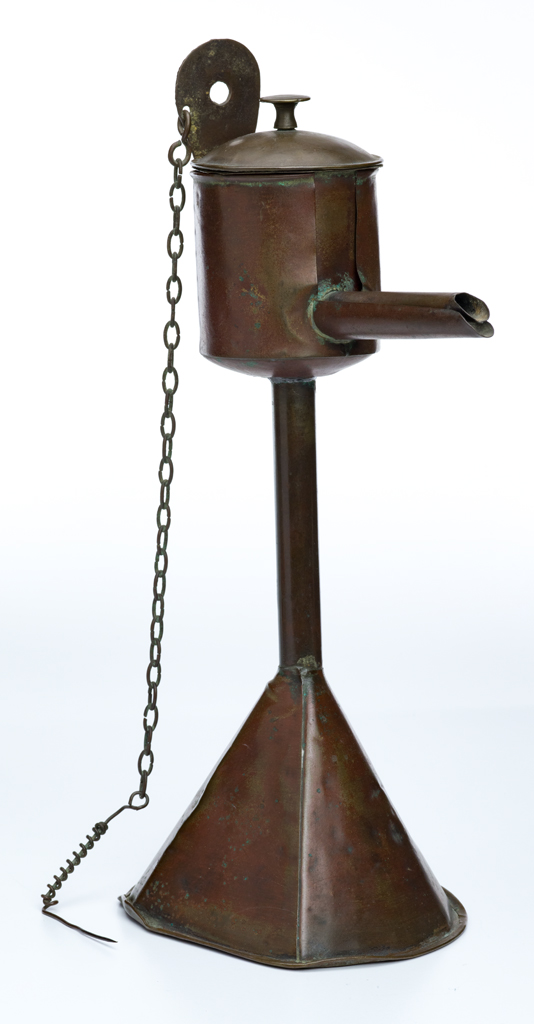 An oil lamp made of metal, has a seperate round central oil container with lid and a spout at one end where the tip of an oil soaked wick could protrude. A pick, for rescuing the wick is on the end of a chain attached at the top of the main base. The base is a funnel with one flattened side.