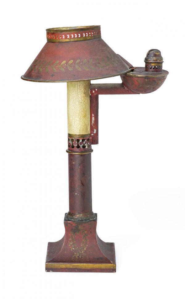 An oil lamp that has been electrified. The shade, oil holder and base has red paint and tole designs. The top of the shade, as well as the bottom of the 