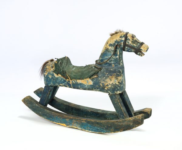 Wooden rocking horse painted blue with a fabric saddle, horse hair mane and tail, and leather bridle and reins. The eyes are nails.