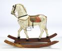 Carved wood, painted white horse. Mounted on red platform with rockers. Tail is binder twine and mane is a synthetic fiber on fiberboard. Reigns and support to stirrip is twill tape, leather ears, harness and saddle, fabric with paper backing for blanket, metal stirrup and misc. hardware