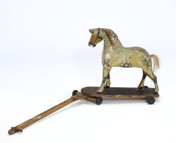 A toy horse pull composed of a carved horse covered in painted gesso with horse hair mane and tail, leather rains, and metal brad eyes. The horse is mounted to a flat board with arch at front on a set of four wheels. At the front is a wooden pull handle.