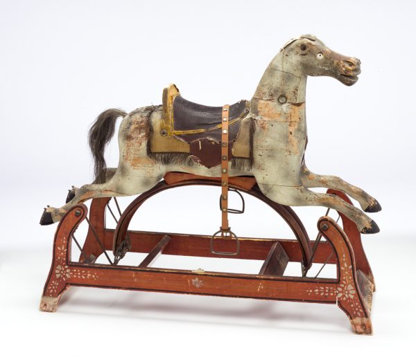 A white rocking horse sitting on top of red stationary base. The horse is made of painted wood, faux-leather, sawdust, horse hair, cloth with fringe, misc. metal fittings, proper left eye is amber colored glass. The horse
moves forward and backward via cast iron components that are mounted to the underside of the horse structure and the outer support frame.