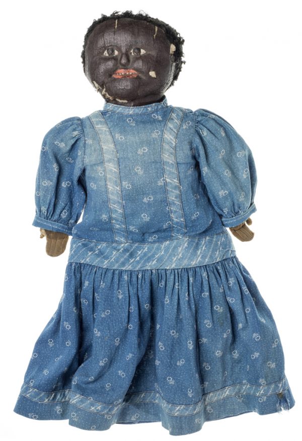 A doll made by hand from cotton padding, fabric, and stockinette. The doll wears a blue patterned cotton dress. Her hair is composed of black yarn and her face and features were painted.
